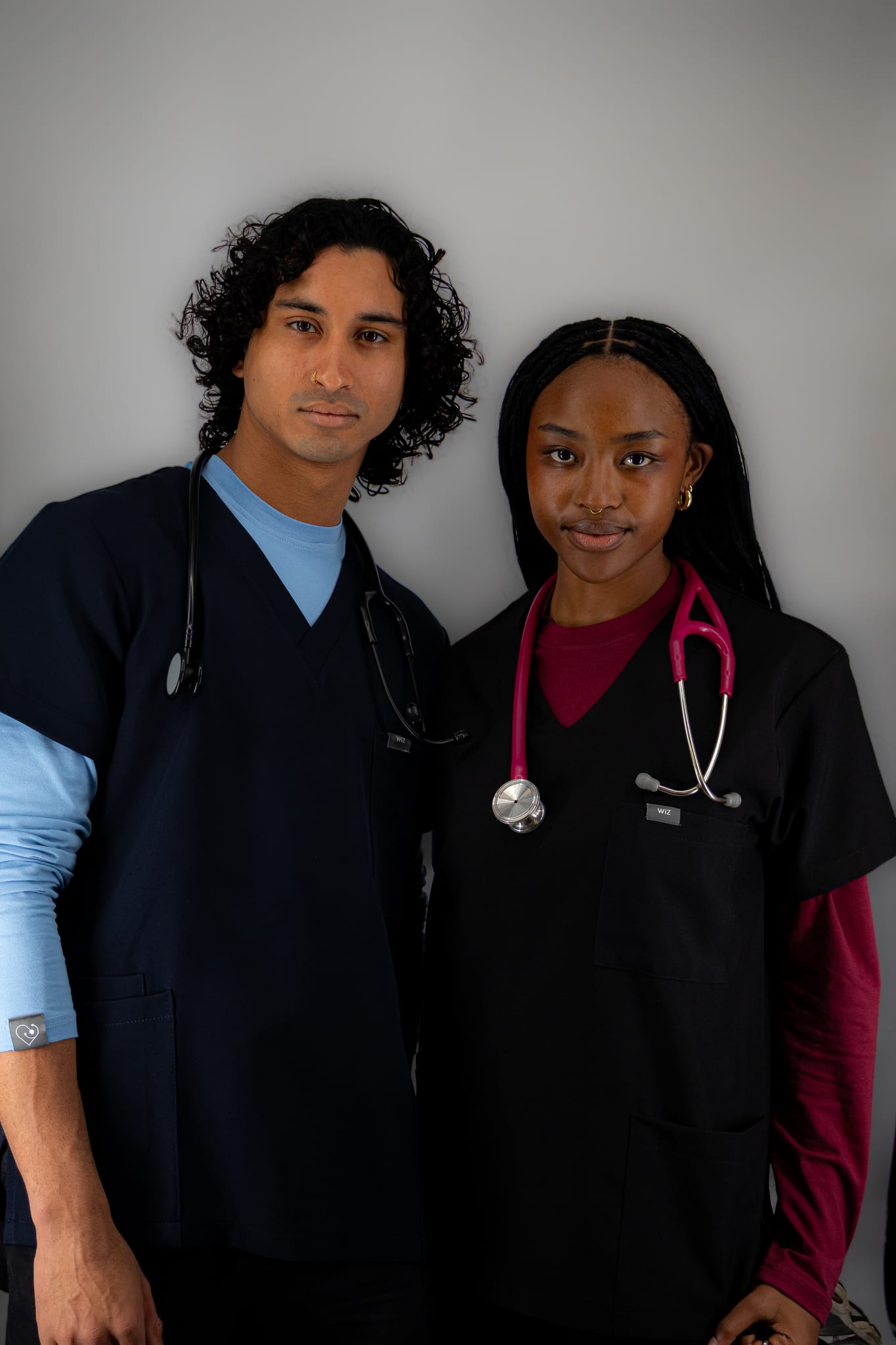 Two students in WiZscrubs: navy scrubs over a sky blue underscrub, and black scrubs over a maroon underscrub